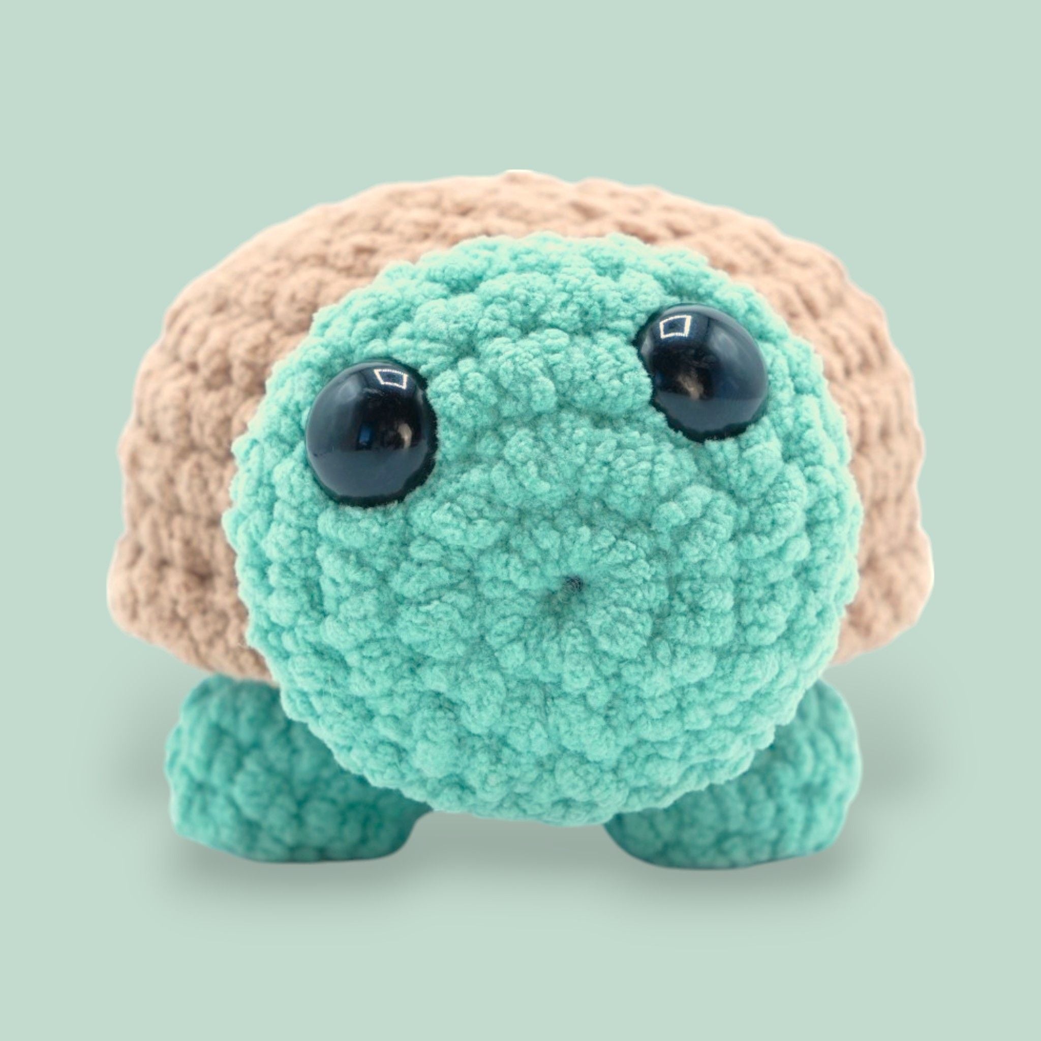 A crochet tortoise plush made from soft chenille yarn in shades of green and brown, with large black eyes and a content expression.