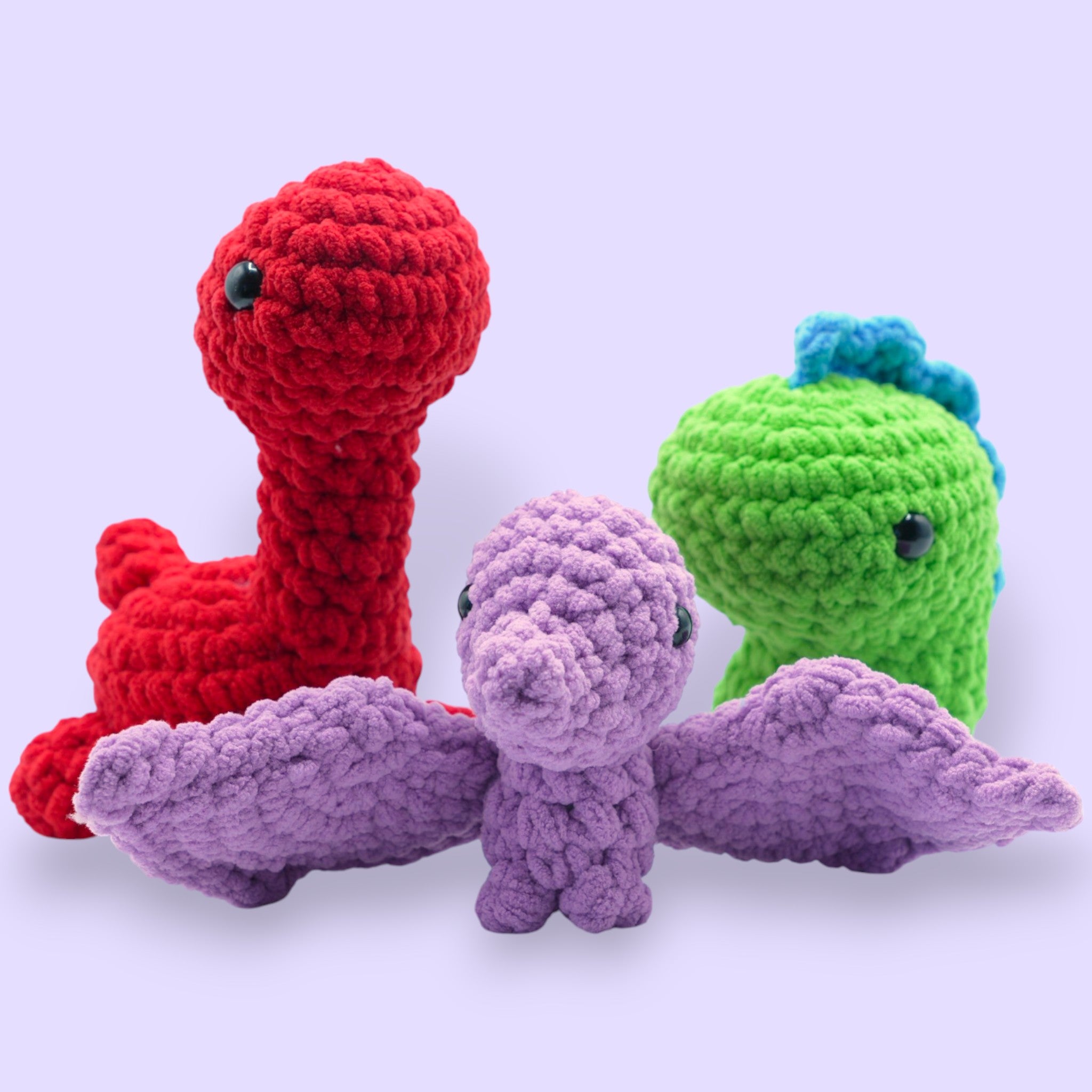 Four crochet dinosaurs in various colors: green T-Rex, red Brontosaurus, and a purple Pterodactyl, all made from soft chenille yarn.