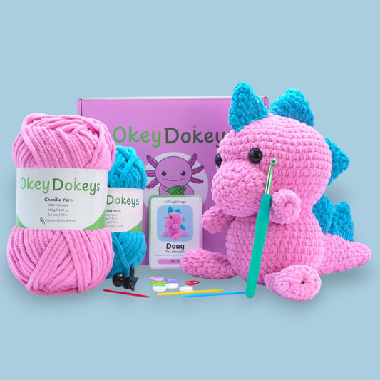 A crochet dinosaur kit displayed with pink and blue chenille yarn, a crochet hook, safety eyes, stuffing needle, stitch markers, and a finished pink dinosaur plush with bouncy spikes.