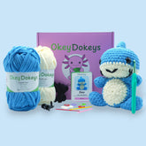 A crochet kit to create a toy shark, with blue and cream chenille yarn, a crochet hook, safety eyes, stuffing needle, and stitch markers, packaged in a box with the product name and an image of the toy shark.
