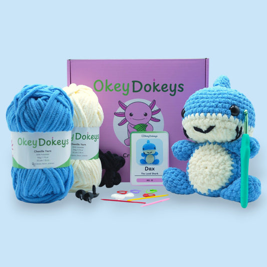 A crochet kit to create a toy shark, with blue and cream chenille yarn, a crochet hook, safety eyes, stuffing needle, and stitch markers, packaged in a box with the product name and an image of the toy shark.