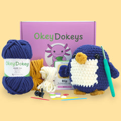A crochet penguin kit by OkeyDokey, featuring yarn in navy blue, white, and orange, alongside a crochet hook, stuffing needle, stitch markers, and a pattern booklet, all arranged on a white background.