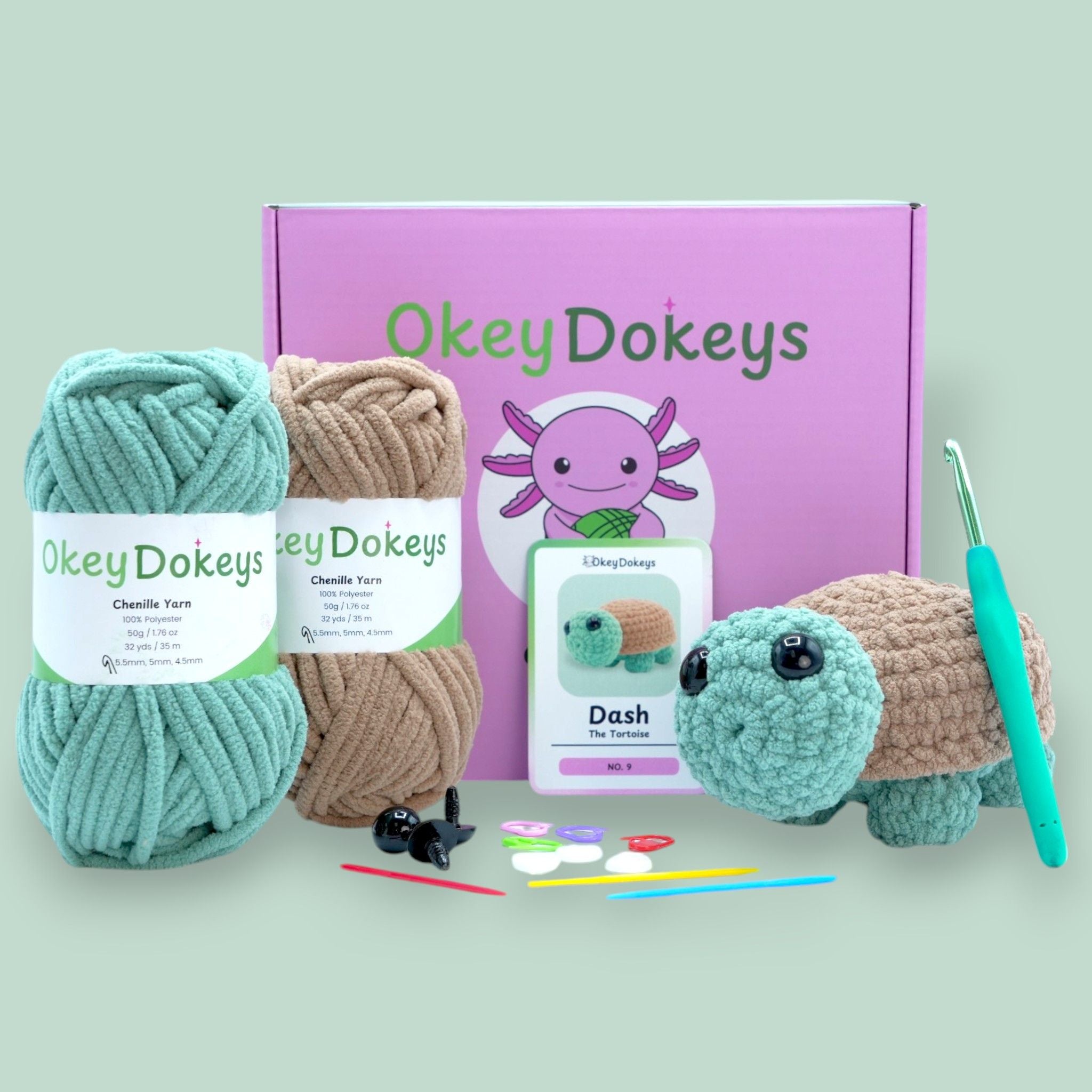 A crochet tortoise kit by OkeyDokeys, featuring yarn in seafoam green and maple syrup tan, a crochet hook, safety eyes, stuffing needle, stitch markers, and a printed tortoise pattern.