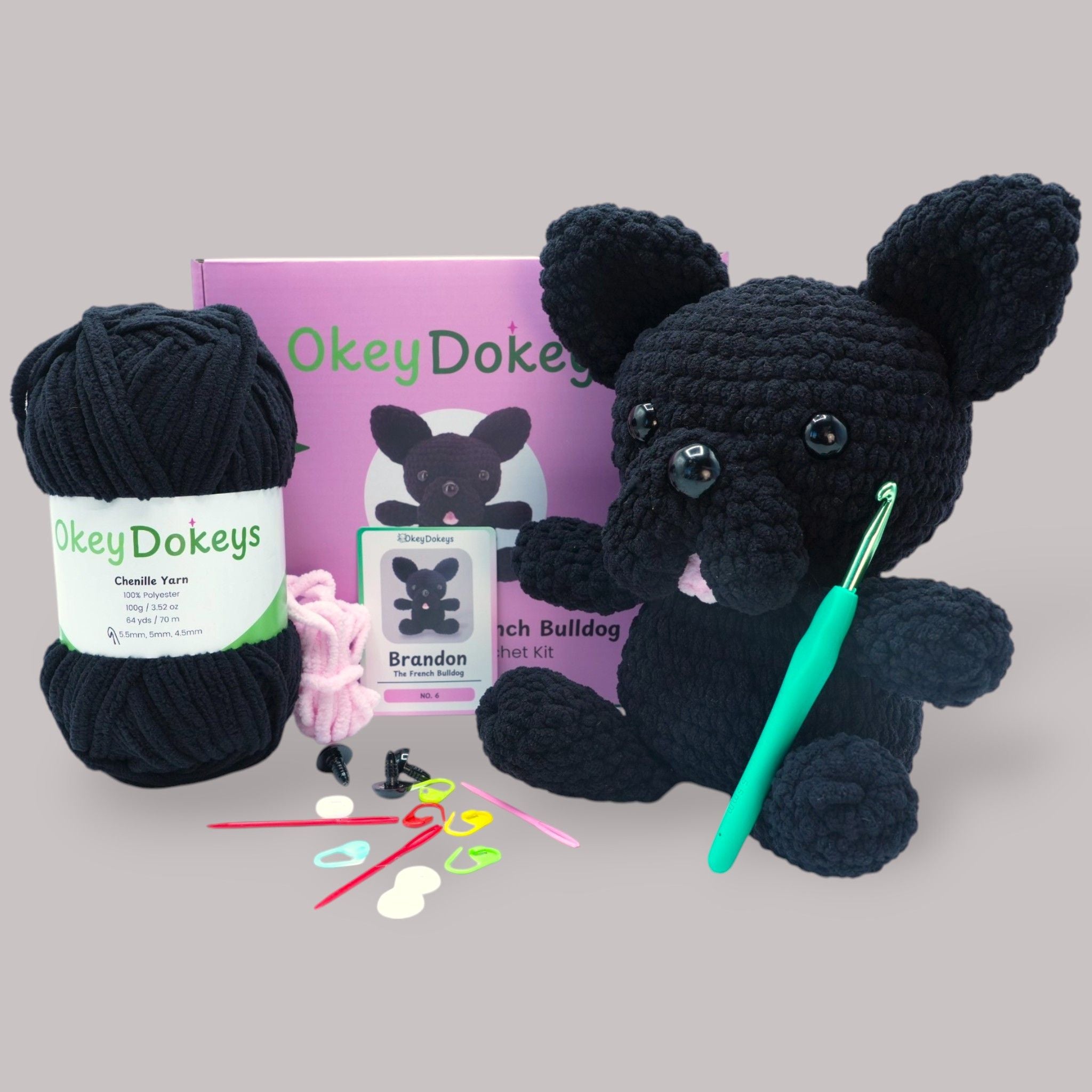 A crochet French Bulldog kit by OkeyDokey, featuring black and dark grey chenille yarn, a crochet hook, safety eyes, stuffing needle, stitch markers, and a pattern booklet.