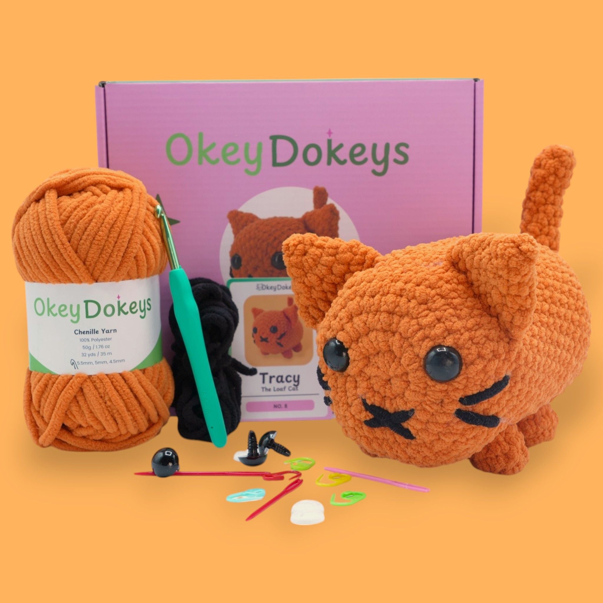 A crochet cat kit by OkeyDokeys, featuring a plush cat toy in orange, pink, and white colors, along with chenille yarn, a crochet hook, safety eyes, stuffing needle, and stitch markers.