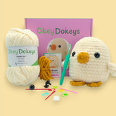 A crochet kit for a baby chicken plushie, featuring a ball of light yellow chenille yarn, a crochet hook, safety eyes, stuffing needle, stitch markers, and the finished plushie chicken displayed in front of a pink box with the brand name 'OkeyDokey'.