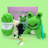 A crochet frog kit by Okey Dokeys, featuring green and white yarn, a crochet hook, safety eyes, stuffing, and other materials laid out on a green background.