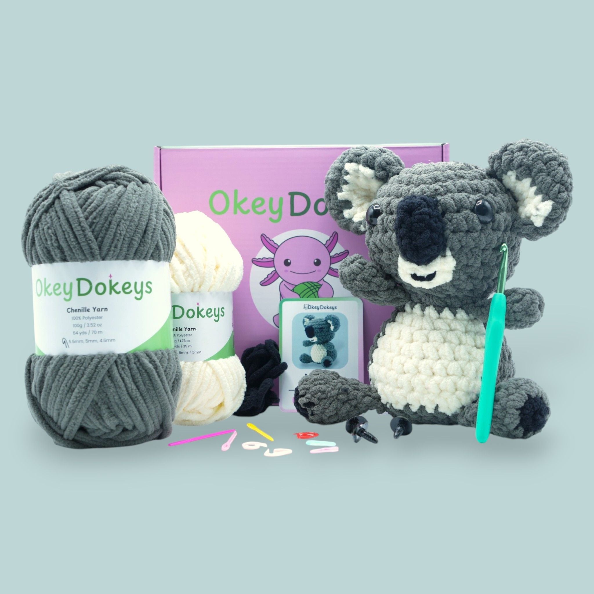 A crochet koala kit with gray, white, and black chenille yarn, a crochet hook, safety eyes, stuffing, scissors, and a pattern booklet, all arranged neatly on a flat surface.