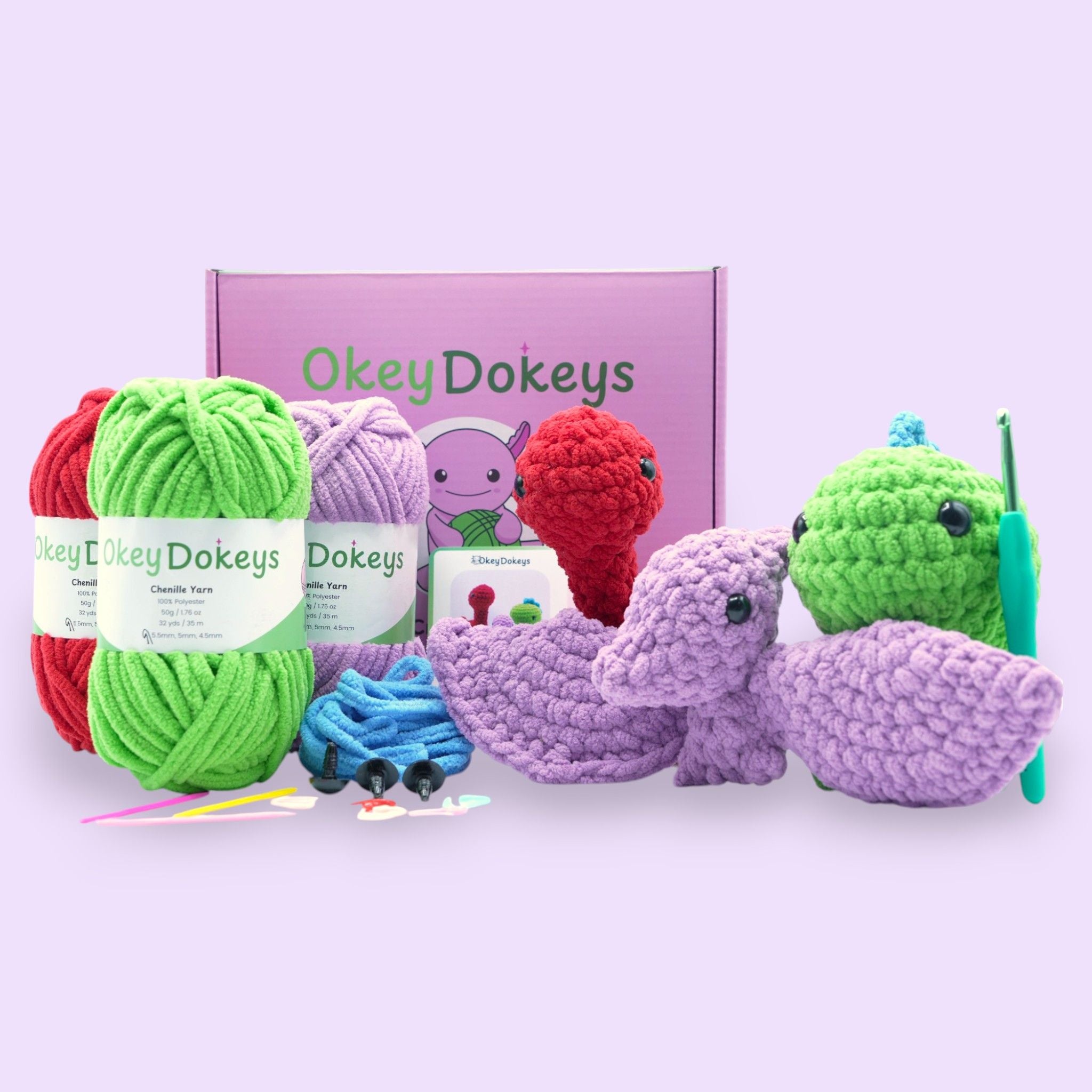 A collection of crochet supplies including yarn in various colors, a crochet hook, scissors, and a box with the brand name Okey Dokeys. There are also three plush crocheted dinosaurs displayed.