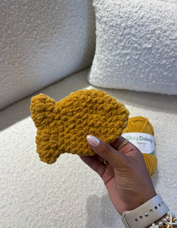 Hand holding a yellow knitted fish-shaped object with a ball of yarn in the background.