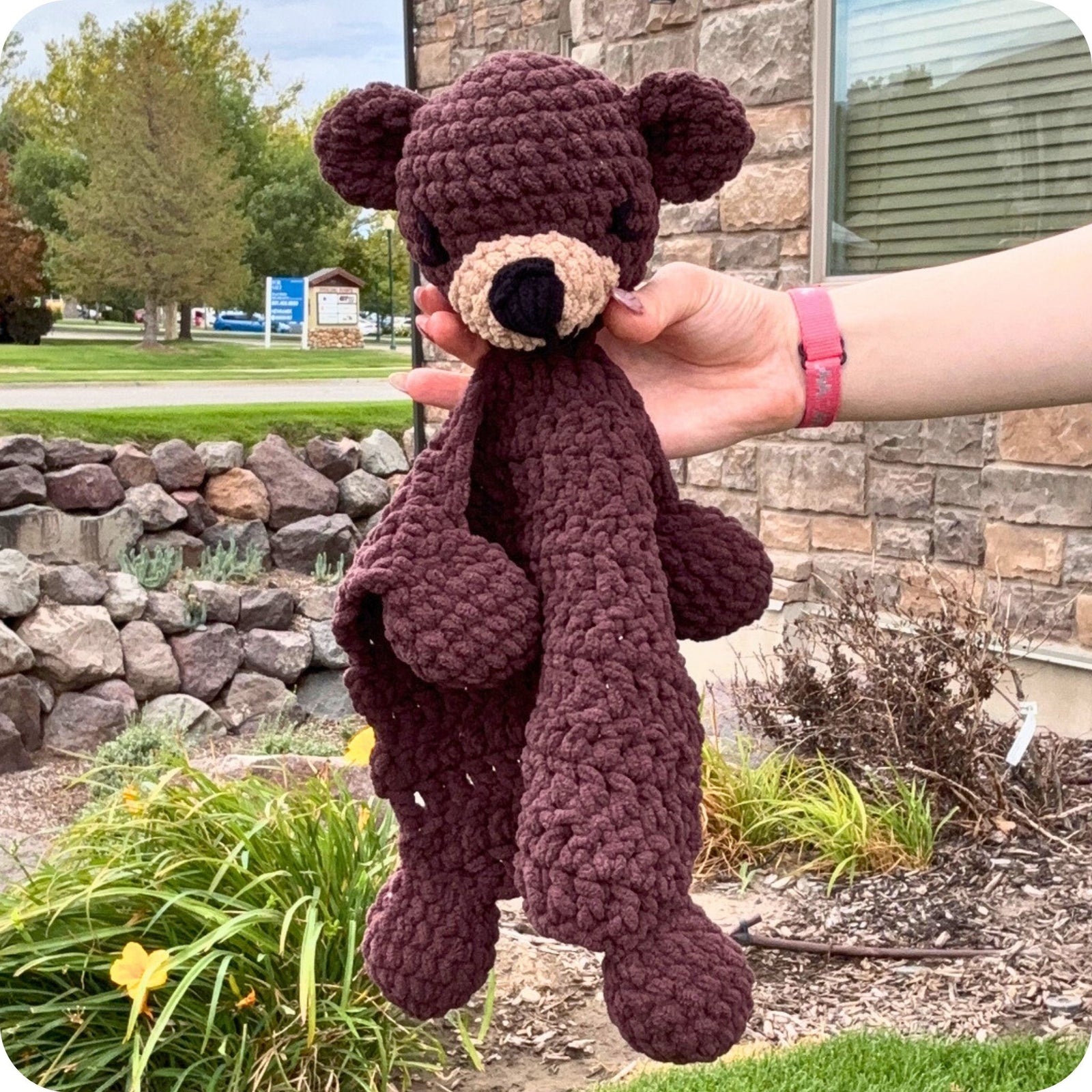 A hand holds a dark brown crocheted teddy bear comforter outdoors in front of a stone wall and greenery. The comforter has a beige stitched snout and nose, black embroidered eyes and nose, and a matching dark brown blanket draped over its body.