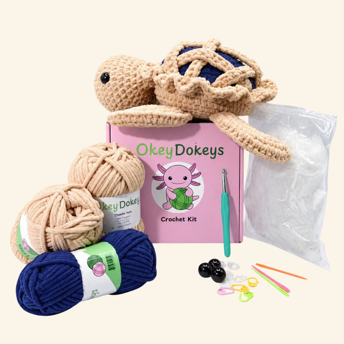 OkeyDokeys crochet kit with a crocheted axolotl toy, two balls of beige yarn, one ball of navy yarn, a pink instruction box, a green crochet hook, and various knitting accessories on a light cream background.