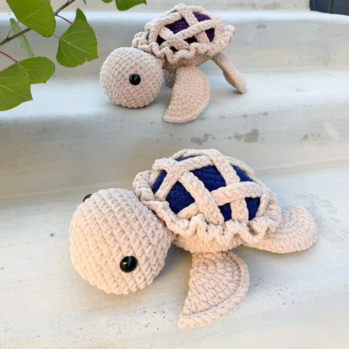 Two handmade crocheted turtle toys displayed on light gray concrete steps, with green leaves in the upper left corner. One turtle is positioned in the foreground and the other is in the background.
