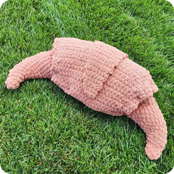 A crocheted plush toy shaped like a crescent moon or horn, in a dusty pink color, lying on a green grass lawn.