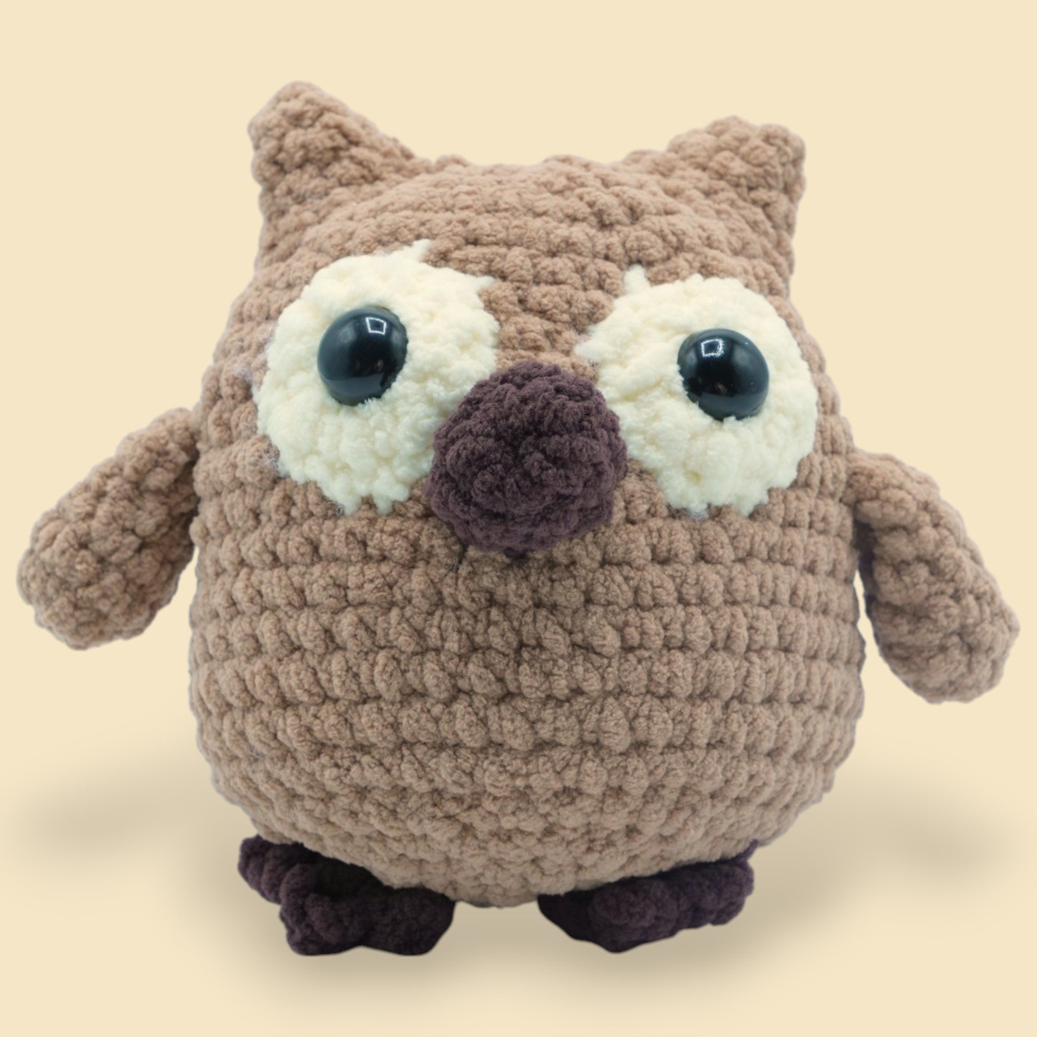 A crochet owl kit with a beige, brown, and white body, black eyes, and a purple perch.