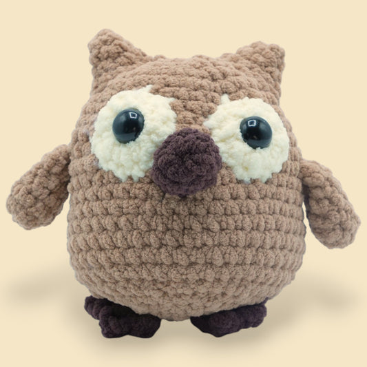 A crochet owl kit with a beige, brown, and white body, black eyes, and a purple perch.