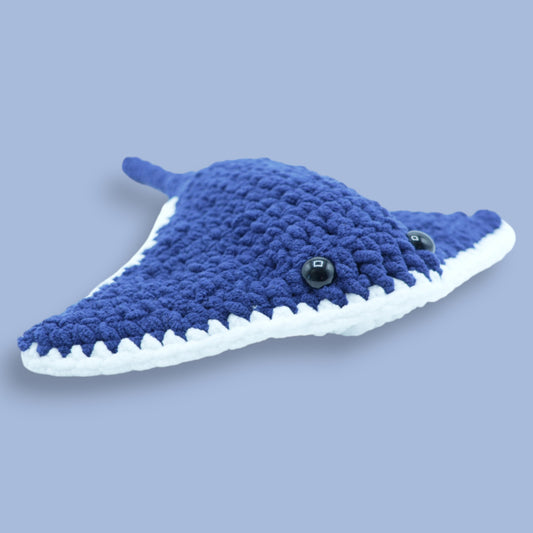 A crochet stingray plush toy made from soft chenille yarn in deep navy and white, with black eyes and a smiling mouth.
