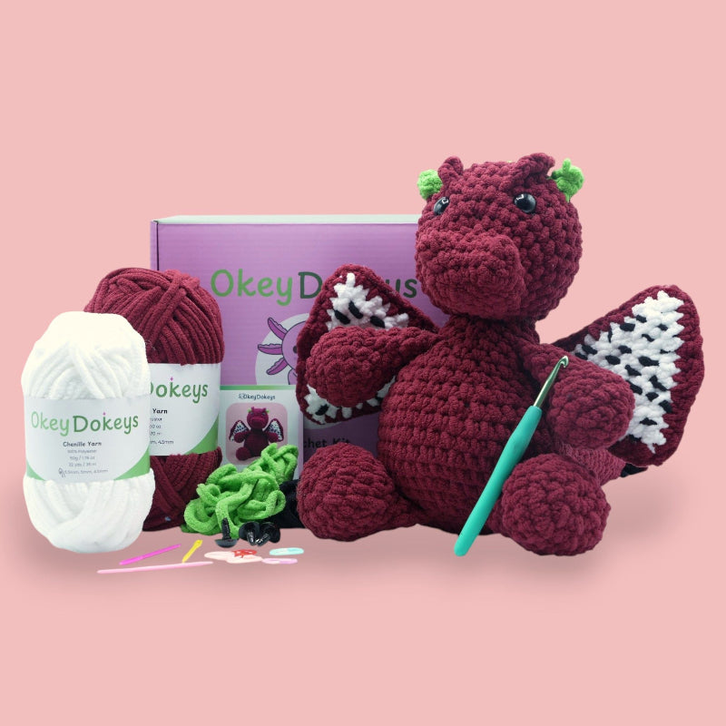 Crocheted dragon toy with yarn, hooks, and packaging on a pink background