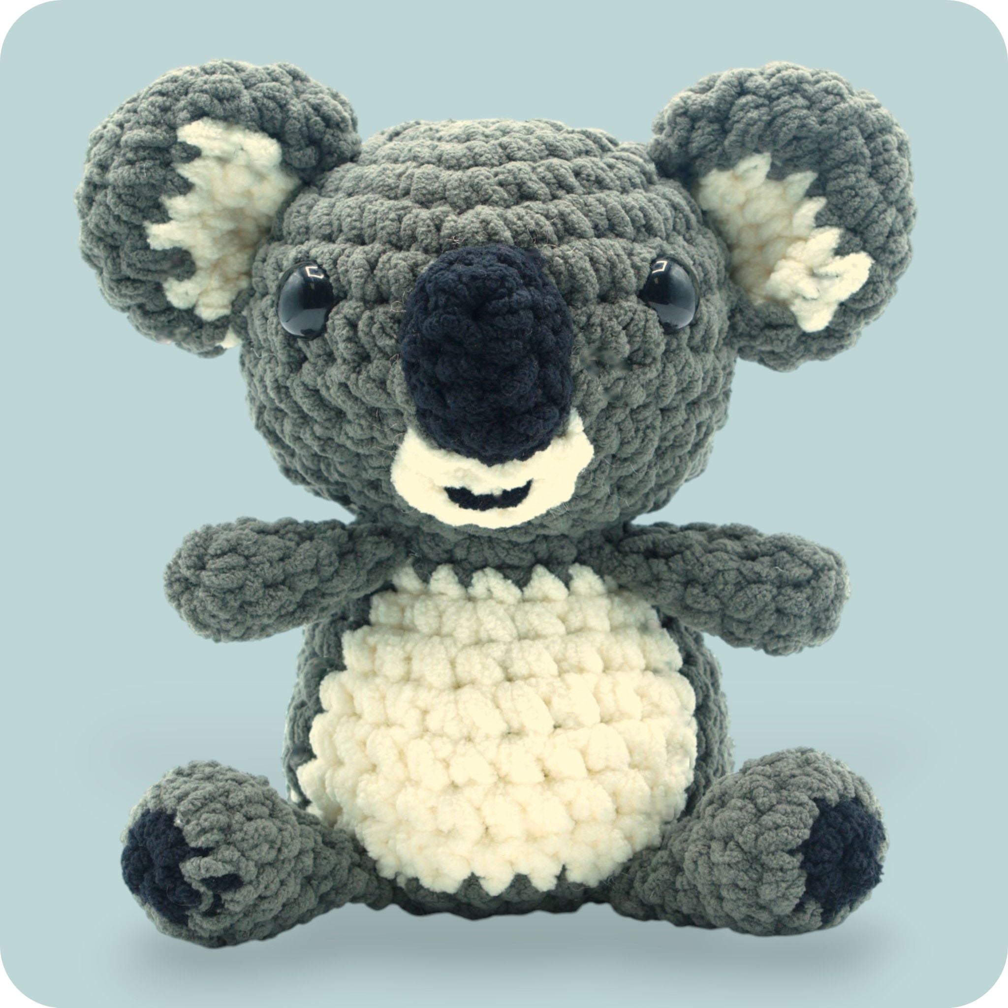 A crochet plush toy resembling a koala, with a gray body, white belly, and black ears.