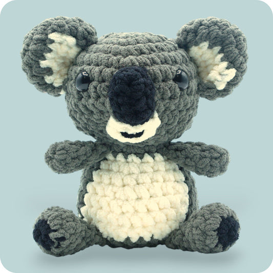 A crochet plush toy resembling a koala, with a gray body, white belly, and black ears.