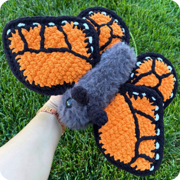A handmade monarch butterfly plush toy held in a person's hand against a green grass background. The plush features bright orange and black textured wings with small light blue accents, a fuzzy gray body, and a small black nose.
