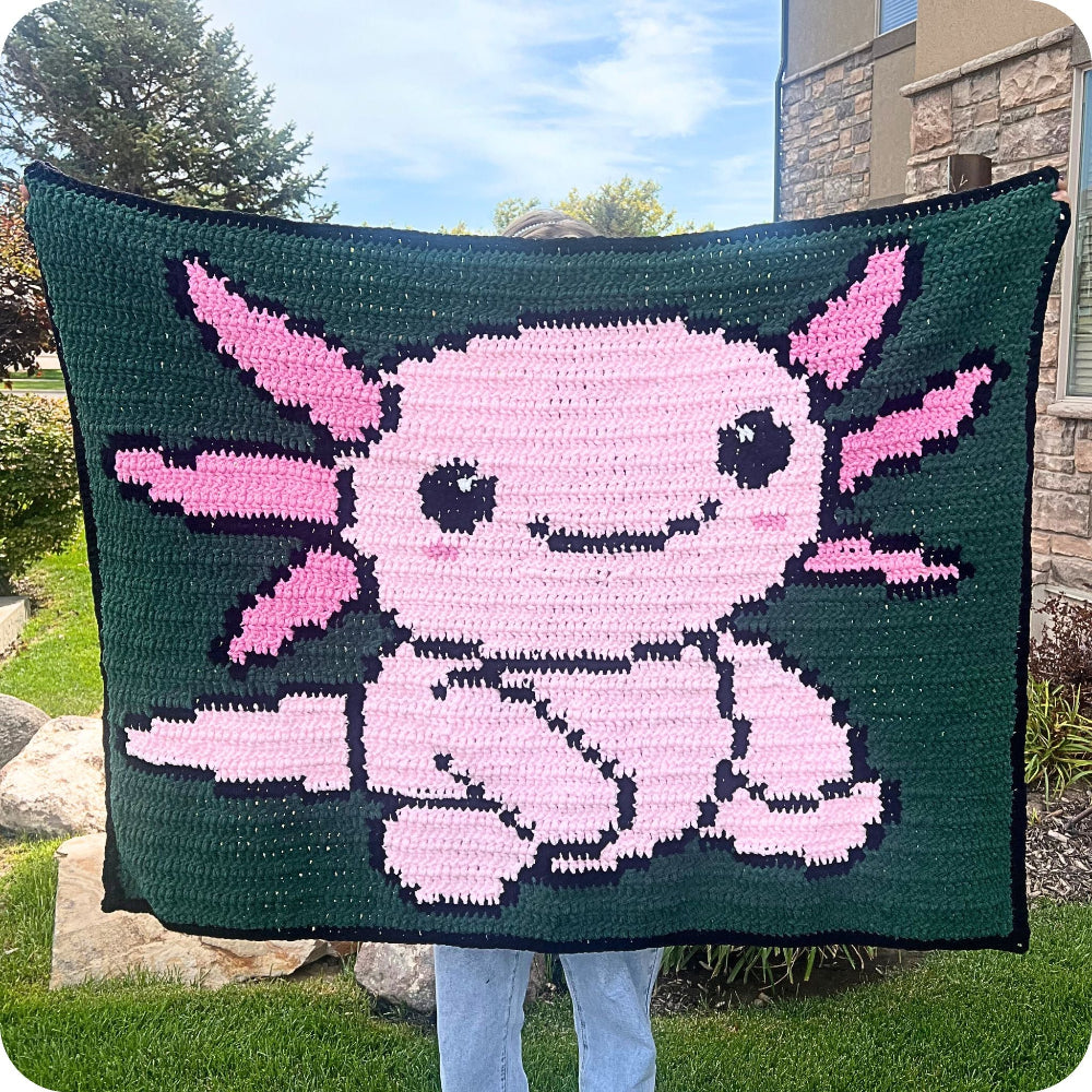Person holding a crocheted blanket with a pink creature design outdoors.