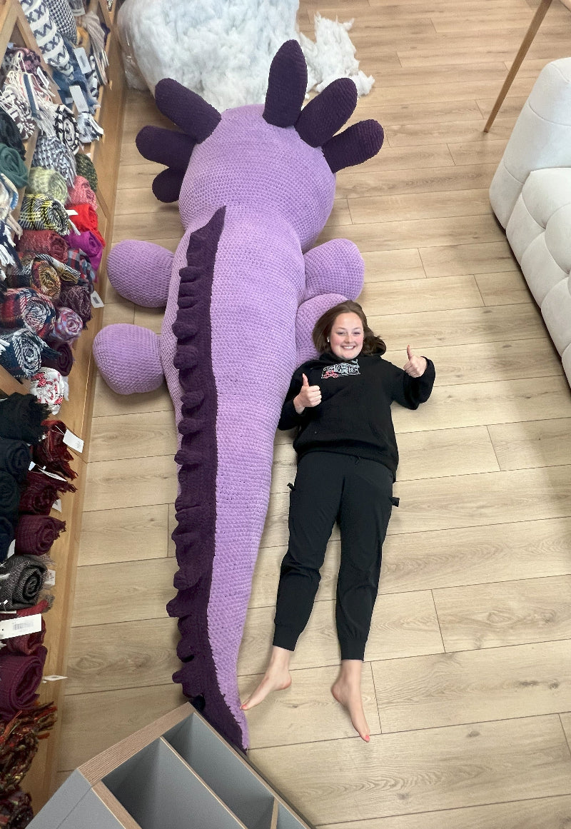 Person lying next to a large purple knitted dragon toy on a wooden floor.