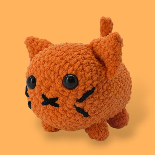 A crochet cat kit by OkeyDokeys, featuring a plush cat toy in orange, pink, and white colors, along with chenille yarn, a crochet hook, safety eyes, stuffing needle, and stitch markers.