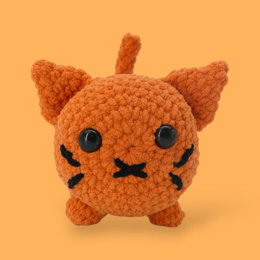 A crochet cat kit by OkeyDokeys, featuring a plush cat toy in orange, pink, and white colors, along with chenille yarn, a crochet hook, safety eyes, stuffing needle, and stitch markers.