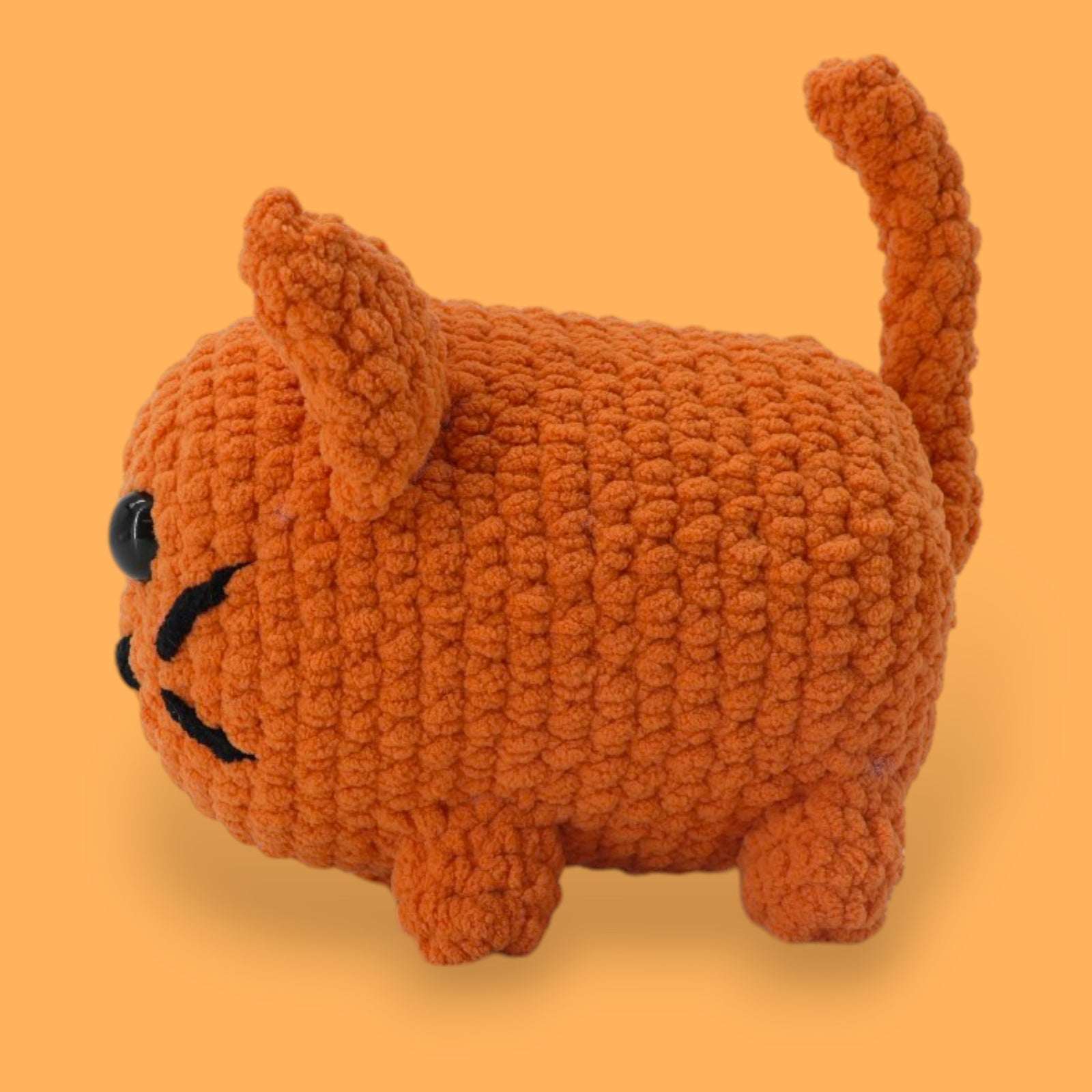 A crochet cat kit by OkeyDokeys, featuring a plush cat toy in orange, pink, and white colors, along with chenille yarn, a crochet hook, safety eyes, stuffing needle, and stitch markers.