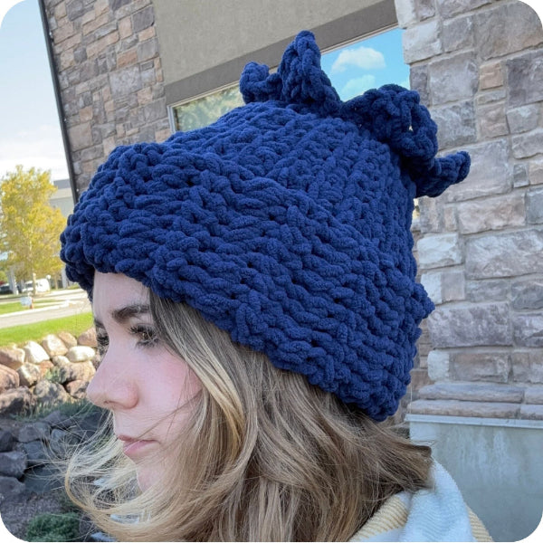 A woman wearing a chunky navy blue knit beanie hat with two knotted pom-poms on top, photographed outdoors in front of a stone building.
