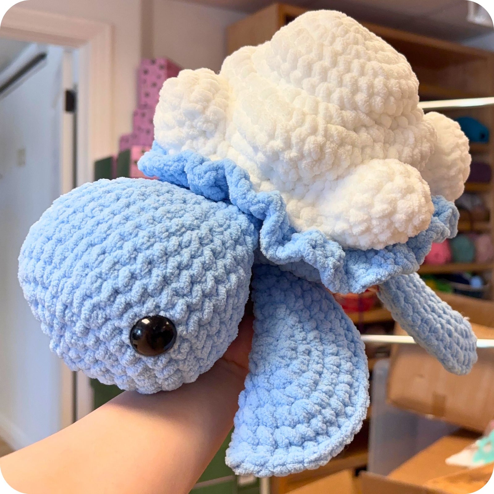 Plush turtle resembling a cloud with blue arms held by a hand indoors.