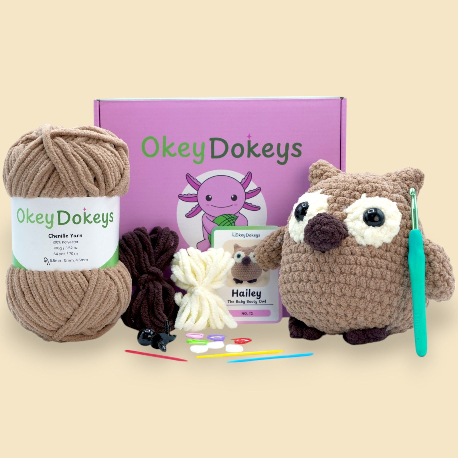 A crochet owl kit by OkeyDokeys, featuring a brown, tan, and white chenille yarn, crochet hook, stuffing needle, and other crafting tools arranged neatly with the finished owl plush.