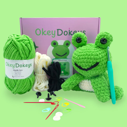 A crochet frog kit by Okey Dokeys, featuring green and white yarn, a crochet hook, safety eyes, stuffing, and other materials laid out on a green background.