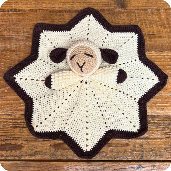 A handmade crocheted baby comforter featuring a sheep head nestled in a star-shaped blanket, displayed flat on a rustic wooden surface.