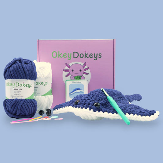 A crochet stingray kit with navy and white chenille yarn, a crochet hook, safety eyes, and a pattern guide, all presented on a blue background.